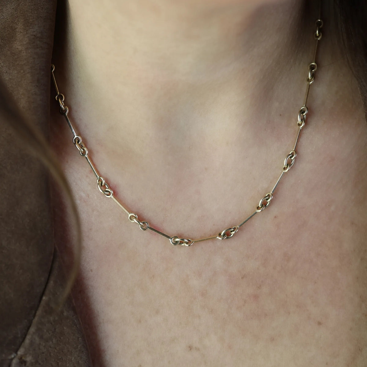 14k yellow gold horsebit chain necklace worn with a brown jacket, its bars and linked rings following the neckline