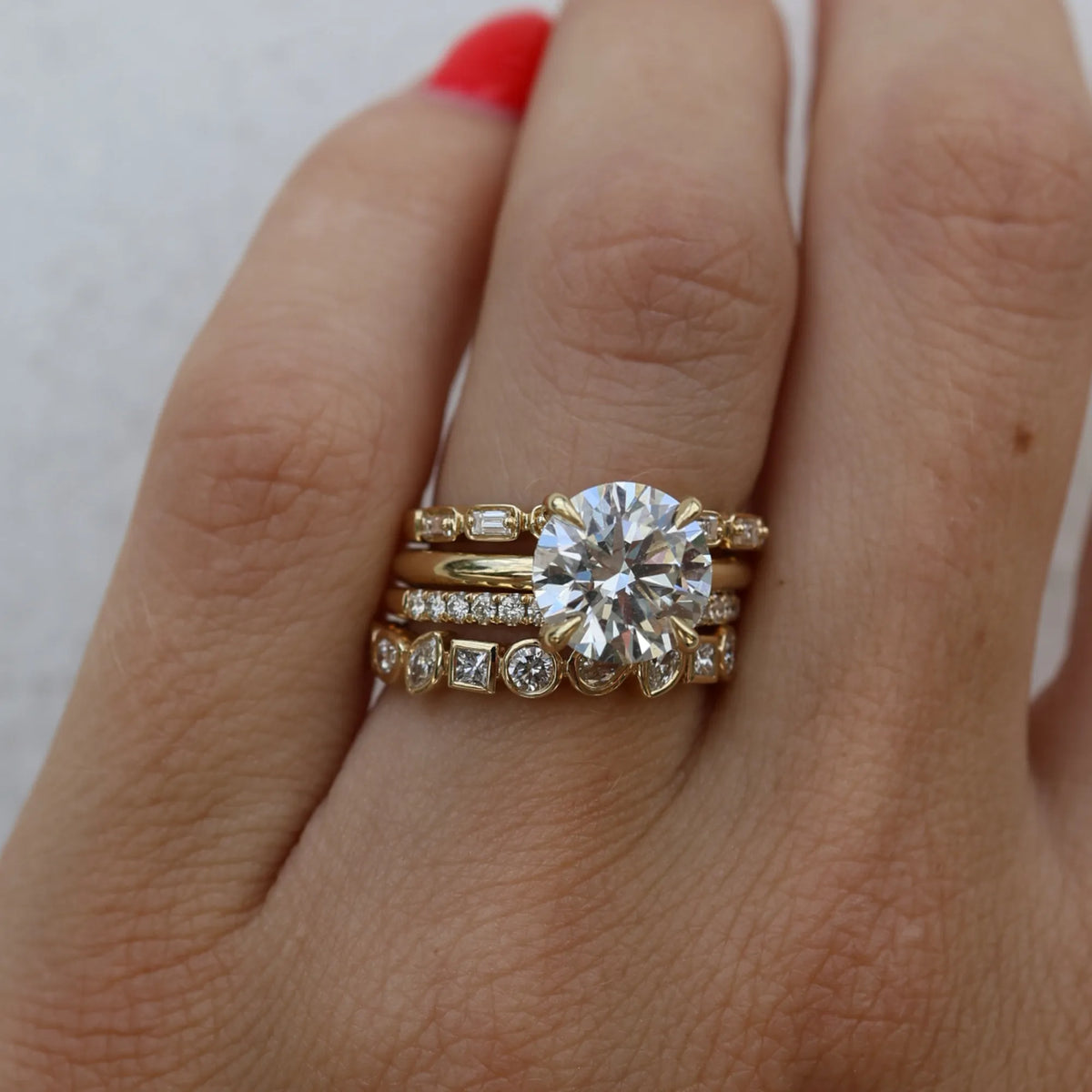 A close-up of a hand wearing a stack of yellow gold rings, featuring a prominent round-cut diamond center stone on the top ring and smaller accent diamonds on the bands below.