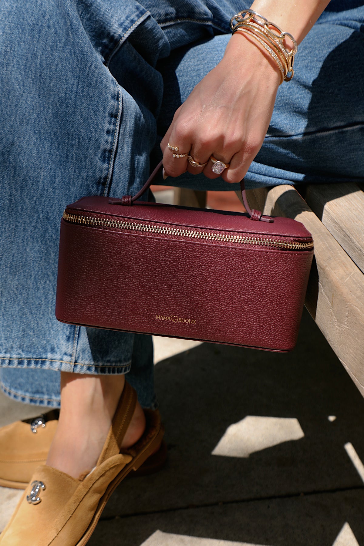 Woman holding the Mama Bijoux luxury jewelry travel case in maroon pebbled genuine leather with gold zipper and carrying handle, styled with fine gold jewelry and denim.