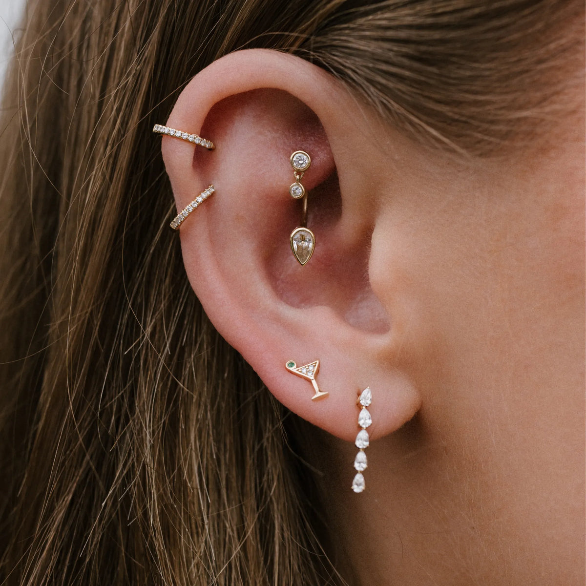 A close-up of a person's ear showcasing a set of gold ear cuff earrings with clear gemstones, including a curved barbell, a small hoop, a teardrop, a cocktail glass, and a dangling drop chain.