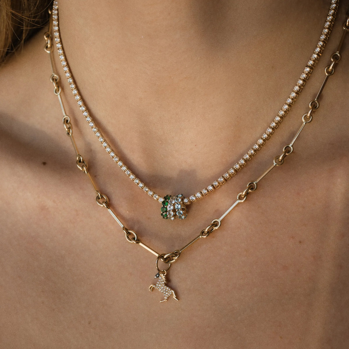 A close-up of a person's neck and upper chest wearing a layered necklace set. The top layer is a delicate strand of small white pearls, and the bottom layer is a gold chain with a small green and clear crystal horse charm and a clear crystal unicorn pendant.