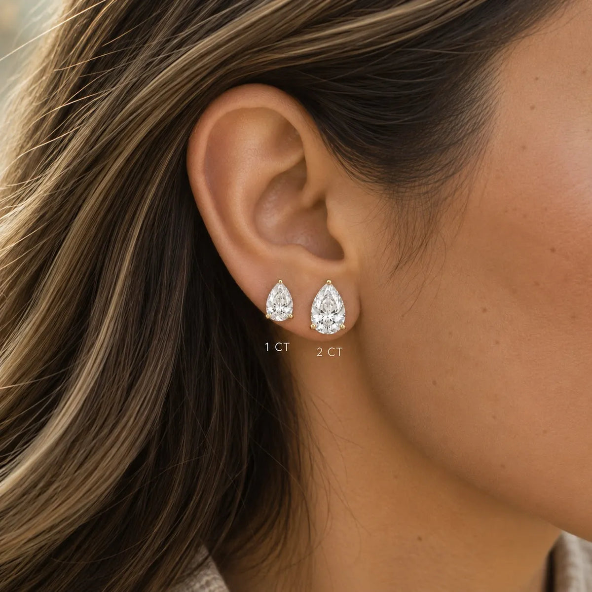 Close-up of a woman's ear wearing two diamond pear shaped stud earrings with clear gemstones, with size markings '1 CT' and '2 CT' near the earrings.