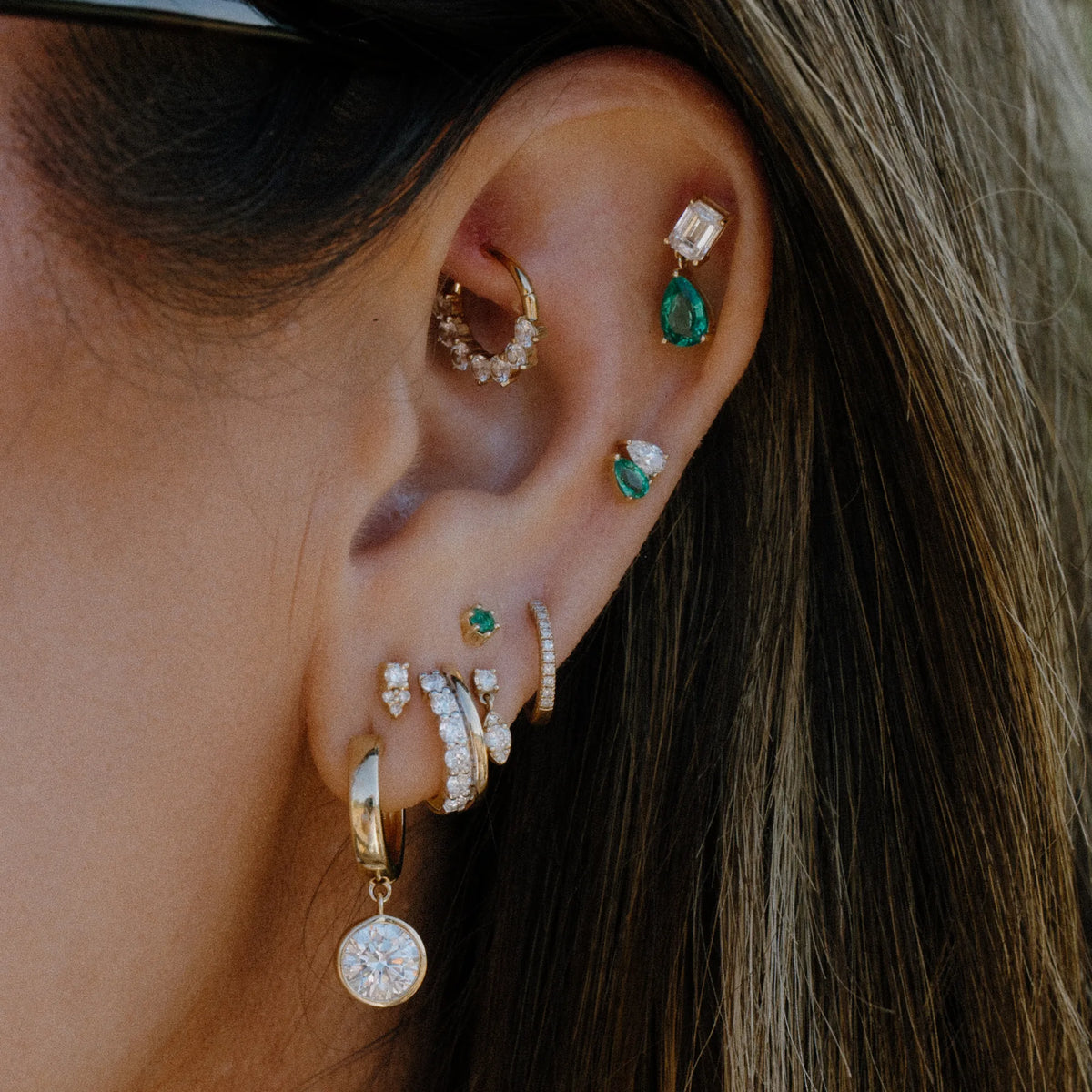 A close-up of a person's ear adorned with multiple gold and silver ear piercings, including hoop, stud, and cartilage earrings with green and clear gemstones.