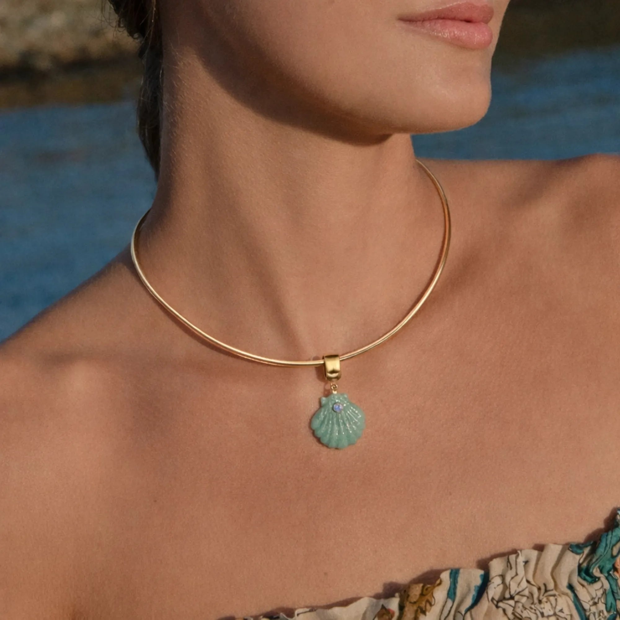Close-up of a person wearing a gold necklace with a turquoise shell pendant by a body of water.