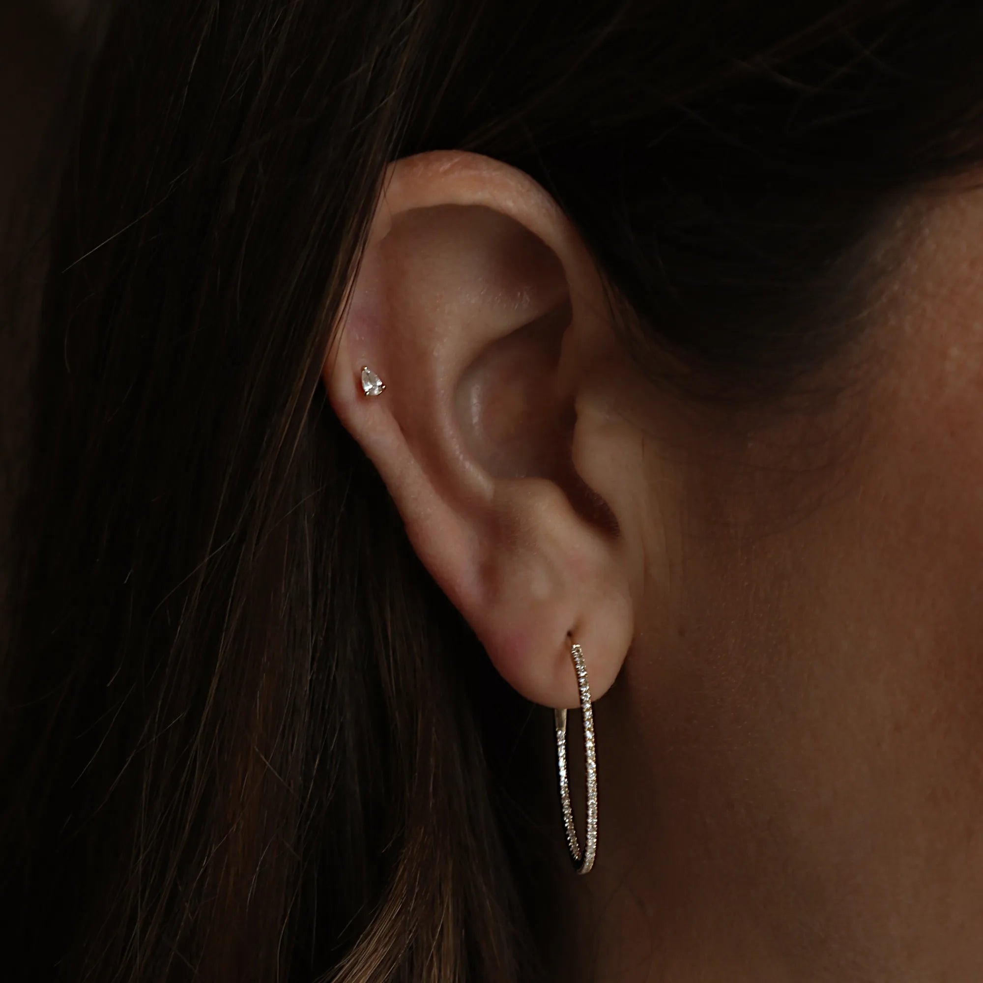 Close-up of an ear wearing a diamond earring with dark hair and skin in the background