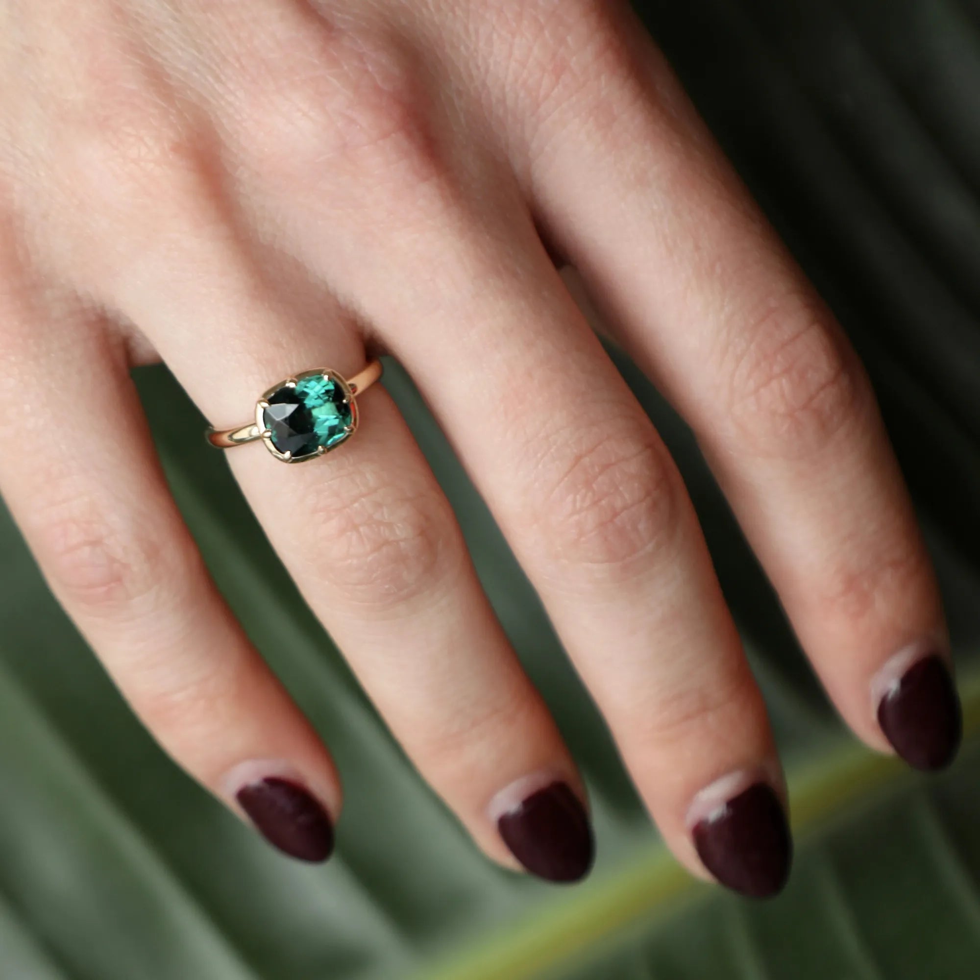 Close-up of a hand wearing a gold ring with a green gemstone on a leafy background