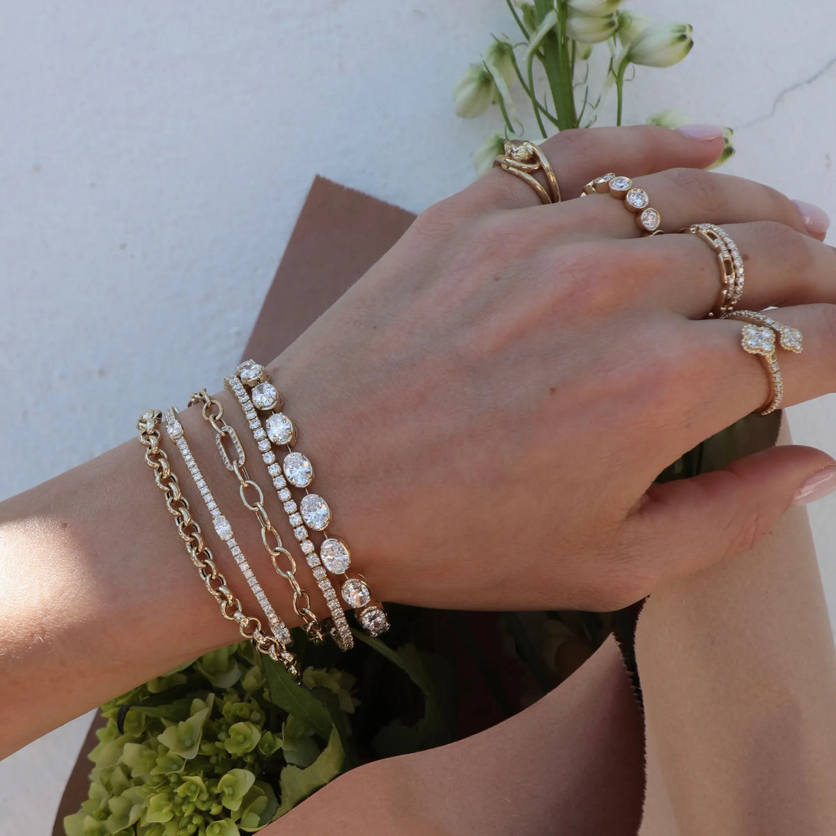 A close-up of a hand wearing multiple gold-toned bracelets and rings with clear stones, posed against a light wall with green flowers in the background.