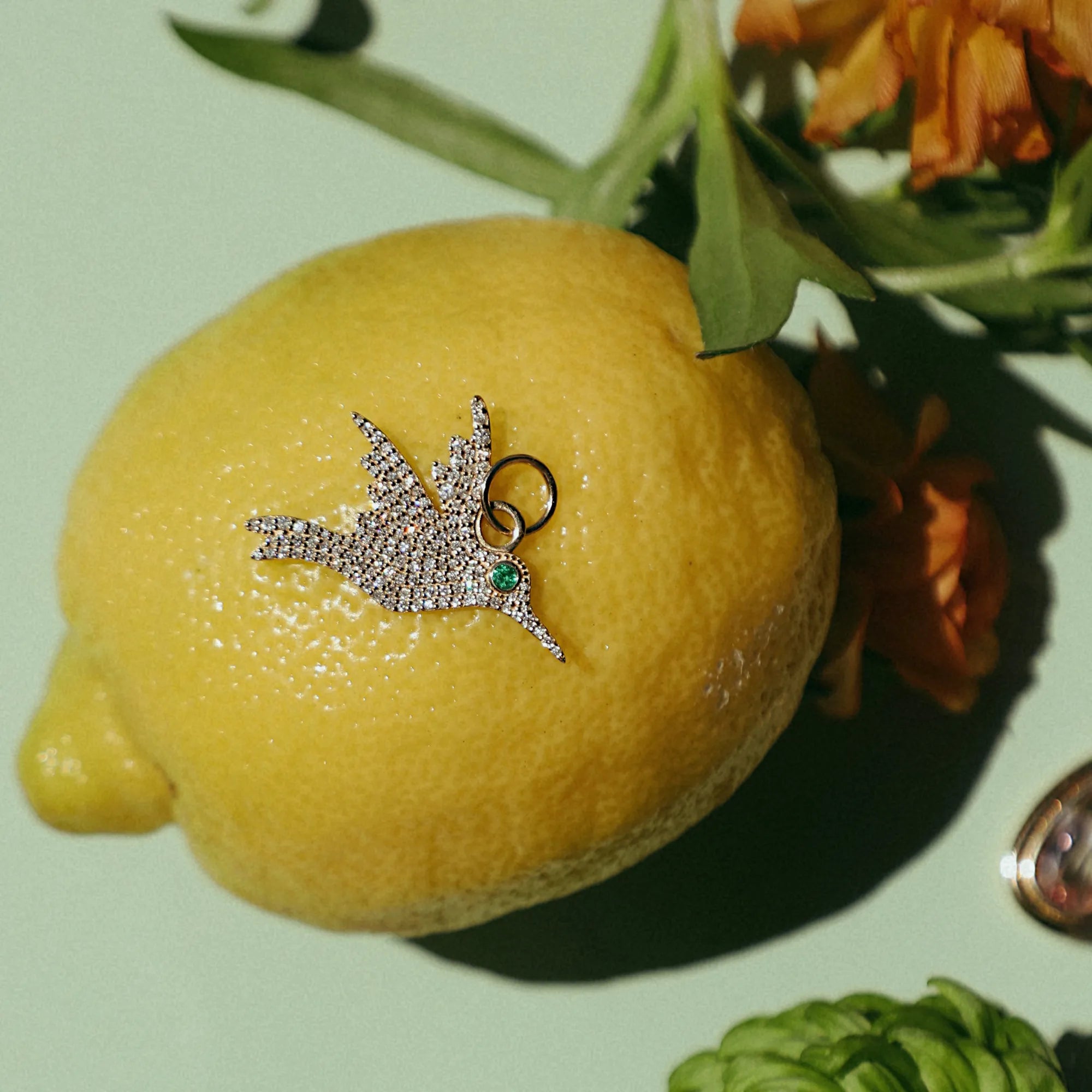 large pavé diamond hummingbird charm in 14k gold with natural emerald eye styled on lemon with flowers