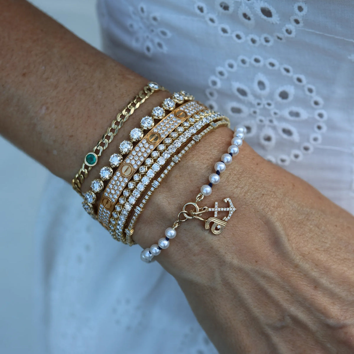 A close-up of a person's wrist wearing a stack of gold bracelets, including chain, pave-set crystal, pearl bead, and anchor charm styles, paired with a white eyelet top.